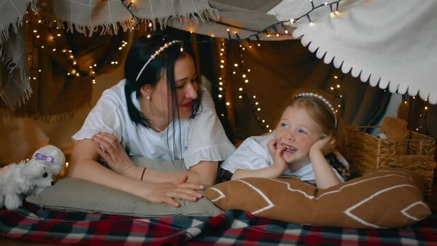 Mom and little daughter communicate while lying in a tent made of blankets and pillows in the bedroom. A family