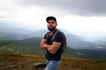 An epic shot of a man with a beard in a cap on top of a mountain. Trekking in the mountains alone, outdoor activities. Concept scene.