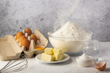 Preparation table for baking, showcasing a bowl of flour, fresh eggs, butter slices, and a whisk on a textured surface.