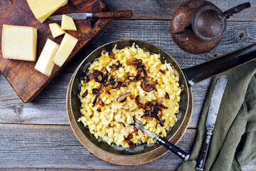 Traditionelle Schweizer Käsespätzle mit gerösteten Zwiebeln serviert als Draufsicht in einer Eisenpfanne auf einem rustikalen Holzbrett in einer Berghütte