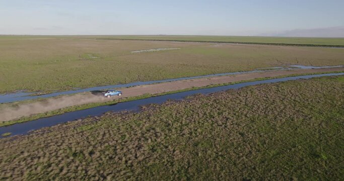 Aerial View Of A Blue Pick Up Truck Driving Very Fast Down The Dirt Road. At The Edge Of The Stream In The Middle Of The Field On A Sunny Day.