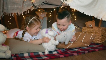 Little boy and girl play with toys and laugh while lying in a tent of blankets and pillows in the bedroom. Children