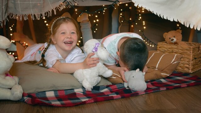 Little boy and girl play with toys and laugh while lying in a tent of blankets and pillows in the bedroom. Children