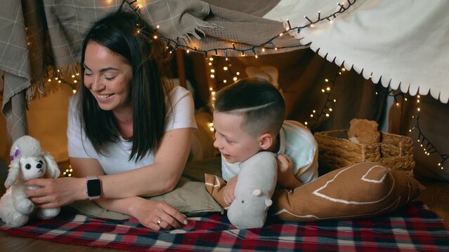Mom and little son play toys while lying in a tent of blankets and pillows in the bedroom. A family