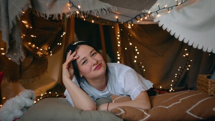 A young woman looks at the camera and laughs while lying in a tent of blankets and pillows in her bedroom. Leisure