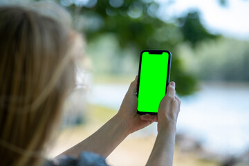 Blonde woman holding phone with empty greenscreen and taking pictures of lake. Mobile photography