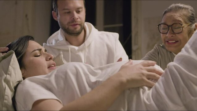 Pregnant woman lying on bed and having contractions at home. Caucasian woman suffering from pain. Asian doula and husband stroking belly and talking with expecting mother. Medicine, pregnancy concept