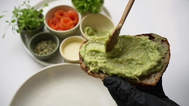 Cook in black rubber gloves spreads guacamole on toast with wooden tablespoon. Ingredients stand on background. Process of cooking closeup