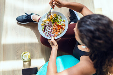 Sporty young woman eating healthy while listening to music sitting on the floor at home. - obrazy, fototapety, plakaty
