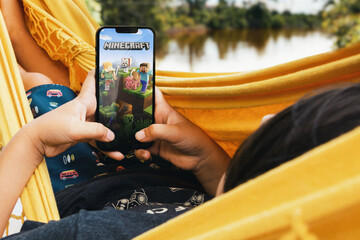 Obraz premium Child lying in hammock with Minecraft mobile game app on smartphone screen. Lake in the background. Rio de Janeiro, RJ, Brazil. May 2022