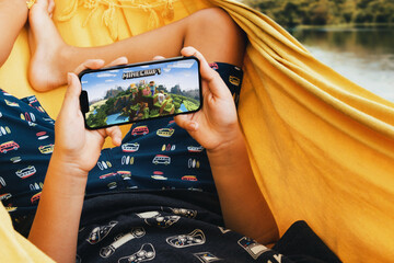 Fototapeta premium Child lying in hammock with Minecraft mobile game app on smartphone screen. Lake in the background. Rio de Janeiro, RJ, Brazil. May 2022