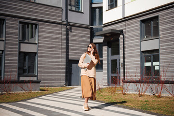 Stylish woman walking on street and using phone, holding a laptop