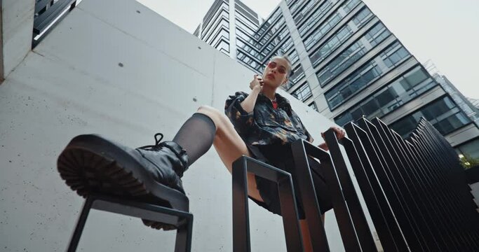 Wide angle urban style sexy young woman wearing colored jacket and cool red glasses standing on stairs leaning leg on railing and looks into camera seductively overlooking high modern buildings