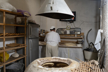 Caucasian man near tandoor in kitchen at bakery. Stone oven for baking tasty bread. Adult man in white clothes getting cook naan using special tool. Baker standing near oven and bakes fresh pastries.
