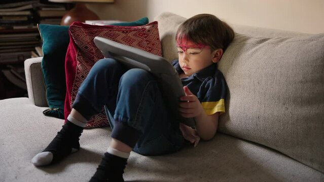 A little boy sitting on couch is watching the cartoon on a tablet with great attention