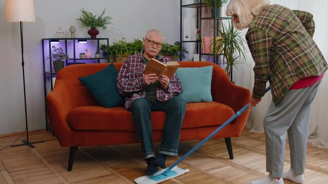 Senior Elderly Grandmother Cleans Wipes Dust On Floor With Mop Under Grandfather Legs While He Reading Book On Couch At Home. Old Mature Family Couple Grandparents Man Woman Doing Housework Routine