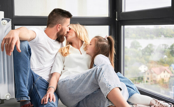 Man and woman sitting on the floor and kissing each other. Happy loving family sitting by the window in newly built apartment. Family, love and new home concept.