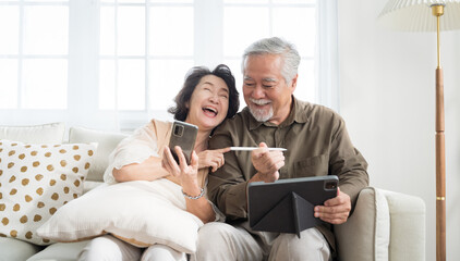 Asian senior couple in living room at home.Wife browsing online on smartphone showing something to her husband while husband is also using a tablet.