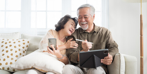 Asian senior couple in living room at home.Wife browsing online on smartphone showing something to her husband while husband is also using a tablet.