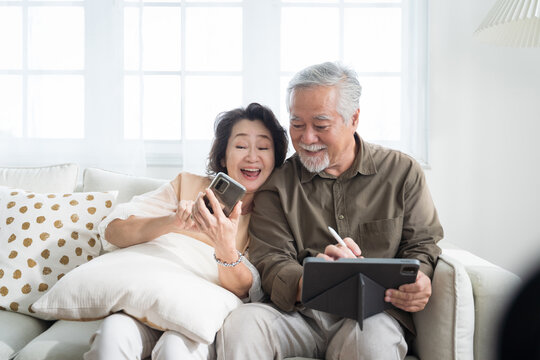 Asian senior couple in living room at home.Wife browsing online on smartphone showing something to her husband while husband is also using a tablet.