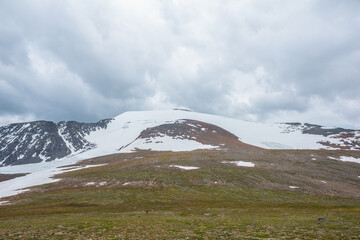 Dramatic landscape with large snowy mountain dome in sunlight under cloudy sky. Colorful scenery with sunlit high snow mountain in dome shape in center at changeable weather. Scenic mountains view.