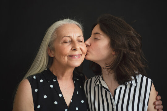 Portrait Of Senior Grandmother With Her Granddaughter Kissing Her Over Black Blackground