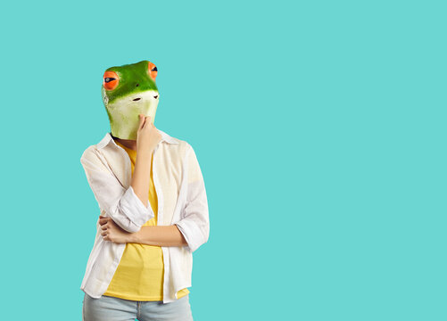 Funny woman in rubber mask of frog stands in pensive pose on turquoise background. Casual woman with frog head stroking chin thinking and looking at copy space. Advertising creative banner.