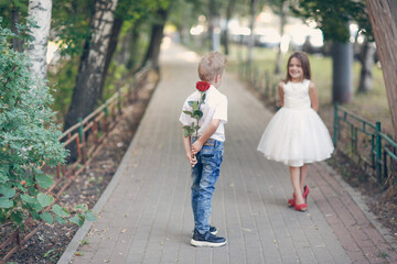 Fototapeta premium cute couple of children boy and girl, boy gives his girlfriend rose for walk in summer. Girl child in high heels, children play dating and courtship, relationship and growing up concept