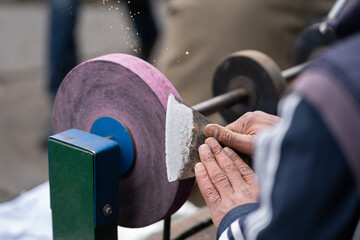 Man holding in hands axe closeup and using sharpening machine. Senior man hands sharpening rusty axe with whetstone. Male working in carpentry workshop and checks sharpness of ax blade. 