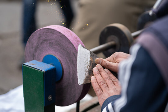 Man holding in hands axe closeup and using sharpening machine. Senior man hands sharpening rusty axe with whetstone. Male working in carpentry workshop and checks sharpness of ax blade. 