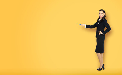 Fototapeta na wymiar Full body length size view of happy excited smiling businesswoman showing pointing advertise at copy space. Confident business woman in black suit standing over yellow color background.