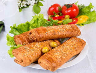 Sweet puff pastry sprinkled with sesame seeds on lettuce leaves, tomato, paprika, green olives on a white dish in light colors