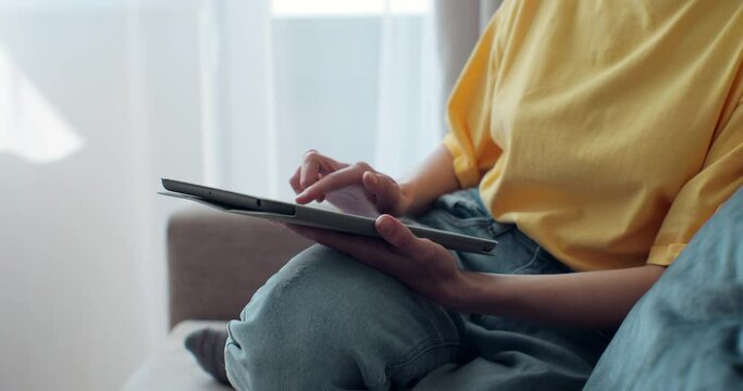 Close up of woman in yellow t-shirt using tablet while sitting on comfortable couch. Female relaxing at home with electronic device.