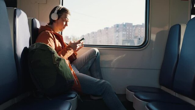 Young man travels alone by electric train with backpack. Man in white wireless headphones scrolls social network pages on smartphone side view