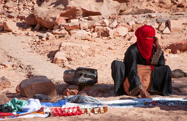 Bedouin woman with covered face sits on sand against rocks in desert selling souvenirs to tourists...