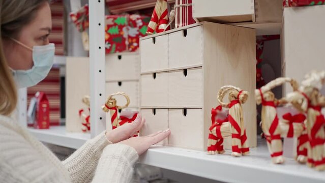 a woman in a protective mask looks at a shelf in a store a wooden box for an advent calendar and a traditional Swedish Christmas goat julbock made of straw