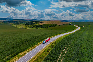 red truck driving on asphalt road along the green fields. beautiful clouds in the sky. Aerial view landscape. drone photography. cargo delivery and transportation concept.