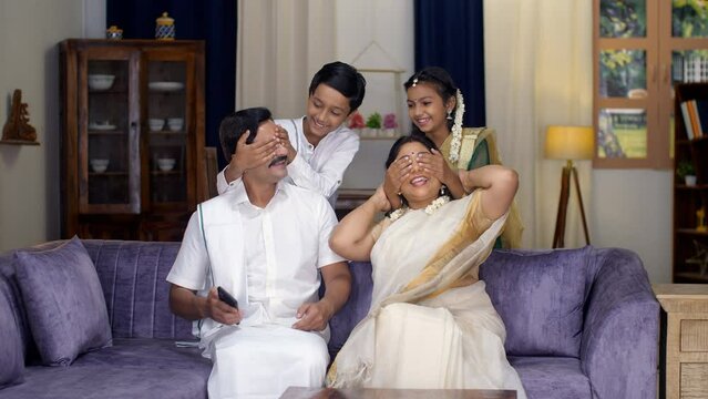 A happy Indian husband-wife in ethnic wear using a smartphone - surprised by kids  South Indian family  traditional dress . Cheerful siblings hugging their parents sitting on a comfortable sofa - a...