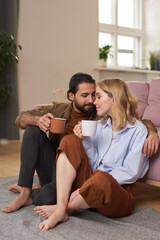 Obraz premium Lovely young heterosexual couple sitting on floor close to each other in living room drinking coffee