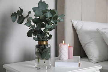 Vase with beautiful eucalyptus branches, book and candles on nightstand in bedroom