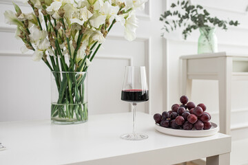 Glass of red wine with grapes and vase on white table