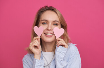 Medium close-up portrait of beautiful young woman with blond hair standing against pink wall background holding paper hearts smiling at camera