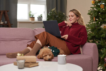 Beautiful young woman wearing warm clothes spending Christmas morning at home relaxing on sofa with her dog in living room watching something in Internet