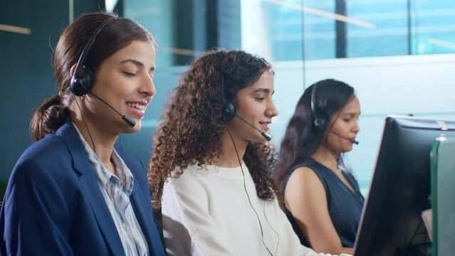 Young modern attractive smiling Indian Asian women or female crew of call center employees wearing headsets or headphones interacting with clients in a corporate office. Customer care support concept.
