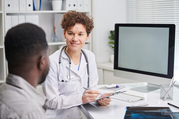 Horizontal shot of modern female doctor wearing white coat sitting in front of patient listening to patients health complaints and writing notes