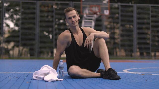 Sporty muscular man sitting on the floor of the basketball court resting before morning run. Young fitness man training on fresh air showing thumb up.
