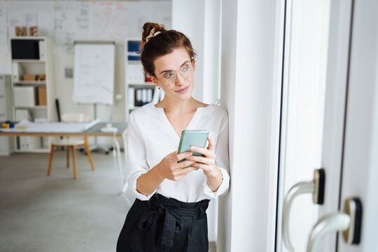 young woman with glasses and smartphone in hand stands in office and looks to the side