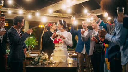 Beautiful Happy Lesbian Couple Celebrate Wedding at an Evening Reception Party with Diverse Multiethnic Friends. Queer Married Couple Standing at a Dinner Table, Kiss and Cut Wedding Cake.