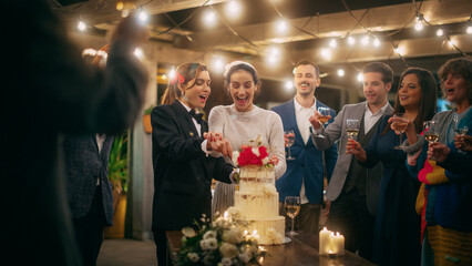 Beautiful Happy Lesbian Couple Celebrate Wedding at an Evening Reception Party with Diverse Multiethnic Friends. Queer Married Couple Standing at a Dinner Table, Kiss and Cut Wedding Cake.