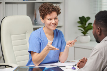 Portrait of successful mature Caucasian female medical specialist sitting at table in front of unrecognizable patient discussing treatment plan